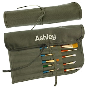 May include: An olive green canvas brush roll, open and closed, with the name "Ashley" printed in white. The open roll displays various paint brushes with colorful handles and different brush head shapes. The closed roll is tied with a matching fabric tie.