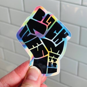 Peut inclure: Autocollant holographique avec un poing noir et l'inscription "Black Lives Matter" en lettres grasses. Le bord de l'autocollant présente un effet irisé arc-en-ciel, créant un design coloré et accrocheur.