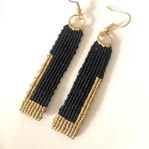 Puede incluir: Un par de pendientes de macramé hechos a mano. Los pendientes presentan un diseño rectangular con un centro negro y bordes dorados. Los pendientes tienen ganchos dorados y se muestran sobre un fondo blanco.