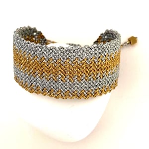 May include: A woven bracelet with a chevron pattern. The bracelet features alternating bands of silver and gold threads. The bracelet is adjustable and has a small gold-coloured square bead at the end of the adjustable cord.
