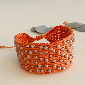 Peut inclure: Bracelet en macramé orange avec des rangées de perles grises. Le bracelet est fait à la main et présente un motif tissé. Le bracelet est de style manchette et possède des liens réglables. Fond crème clair.