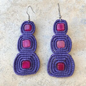 May include: A pair of handmade purple macrame earrings with three stacked, concentric circles. Each circle features a square, pink gemstone. The earrings have silver-colored ear wires.