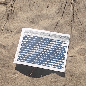 May include: A white and blue 2026 moon phase calendar with detailed illustrations. The calendar is lying on a sandy surface, with the text "2026" at the top. The calendar features a series of circles and squares.
