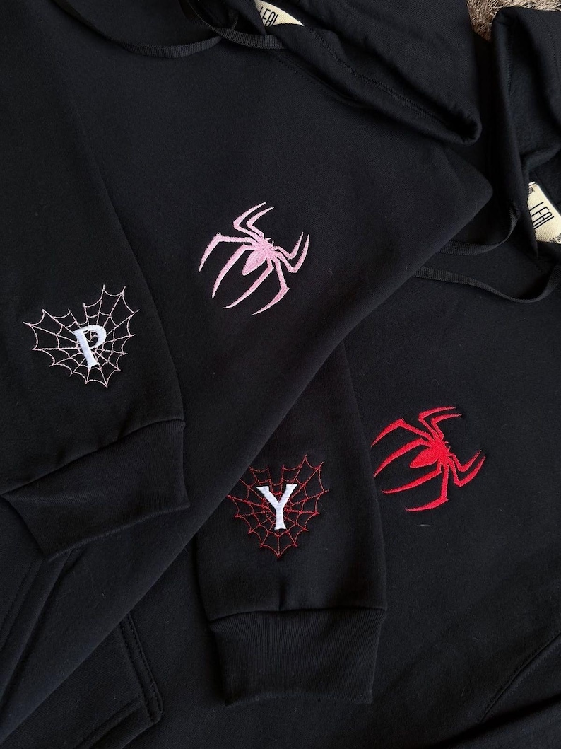 May include: Black hoodies with embroidered spider and spiderweb designs. One hoodie features a pink spider, another a red spider, and the third has a white letter "Y" in a red spiderweb. The fourth hoodie has a pink spider and a white letter "P" in a spiderweb.