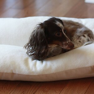 May include: A small, cream-colored dog bed with a light brown trim. A brown and white dachshund is curled up on the bed. The bed has a soft, plush appearance. A small pink tag with the brand name is visible.