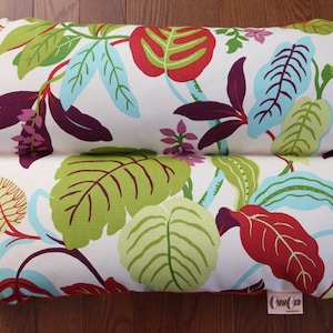 May include: A white pillow with a floral pattern featuring green, red, purple, and blue leaves. The pillow has a red border and a small orange label with the text "Cunard" on it.