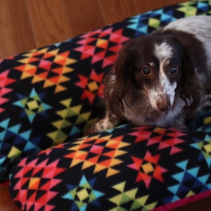 May include: A small, brown and white dog with brown eyes is lying on a colorful, geometric patterned dog bed. The bed is made of a soft, plush fabric and has a black background with bright red, yellow, blue, and green geometric shapes.
