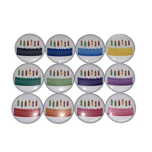 May include: Twelve 1-inch round buttons with a birthday cake design. Each button features a white cake with lit candles and a different colored frosting: black, blue, yellow, purple, green, red, orange, pink, teal, light blue, dark green, and light purple.