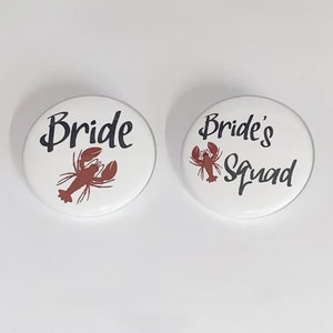 May include: Two white round buttons with black text and a red lobster design. The first button says "Bride" and the second button says "Bride's Squad".