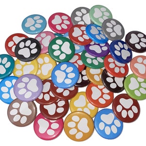 May include: A collection of colorful round buttons with a white paw print design. The buttons are arranged in a pile, showing a variety of colors including red, blue, green, yellow, orange, purple, and pink.