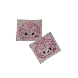 May include: Two gray felt patches with pink and black embroidered brain designs. Each brain has a smiling face with pink hearts for eyes.