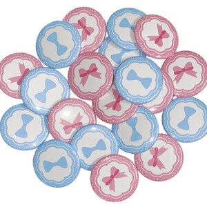 May include: A collection of round, blue and pink buttons with a bow design. The buttons have a white background with a blue or pink bow in the center. The bows are surrounded by a white dotted border.