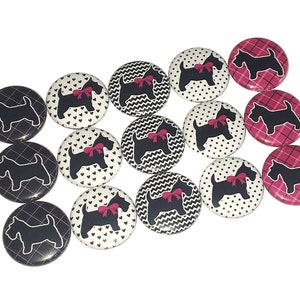 May include: A collection of 14 round black and white buttons with a silhouette of a Scottish terrier wearing a pink bow. Some buttons have a black and white polka dot pattern, some have a black and white chevron pattern, and some have a black and white heart pattern. There are also three buttons with a pink and black plaid pattern.