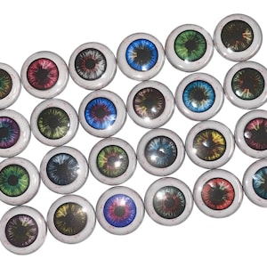 May include: A collection of 25 round buttons featuring colorful eye designs. Each button has a white background with a black outline and a variety of eye colors, including blue, green, red, yellow, and purple.