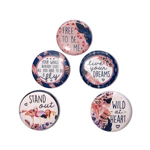 May include: Five round decorative buttons with inspirational quotes and bohemian designs. The buttons feature text like "Free to be me" and "Wild at heart", along with illustrations of feathers and flamingos. The color palette includes pink, blue, and white.