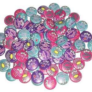 May include: A collection of colorful round buttons with various designs and text related to the game Bunco. The buttons feature phrases like "Let the Good Times Roll!" and "That's How I Roll!" along with images of crowns and dice.