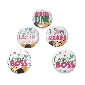 May include: Five white round buttons with text and cookie graphics. The buttons say "Cookie Time," "Just a girl building her empire," "I love cookies," and two say "Cookie Boss." The text is in pink and green.