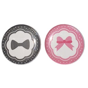 May include: Two round buttons with white polka dots. The button on the left is gray with a white bow tie inside a white scalloped circle. The button on the right is pink with a white bow inside a white scalloped circle.