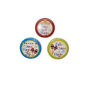 May include: Three round buttons with different designs celebrating Cinco de Mayo. The first button is red with a sombrero and the text "Happy Cinco de Mayo". The second button is blue with maracas and the text "Cinco de Mayo". The third button is green with maracas and the text "Feliz Cinco de Mayo".