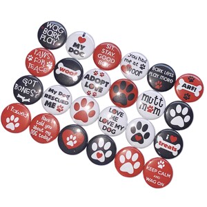 May include: A collection of 20 round buttons with various dog-themed designs. The buttons are black, white, and red with paw prints, hearts, and text. Some of the text includes "Wag Bark Play", "I Love My Dog", "Adopt Love", "Mutt Mom", "Keep Calm and Wag On", and "I Pawed".
