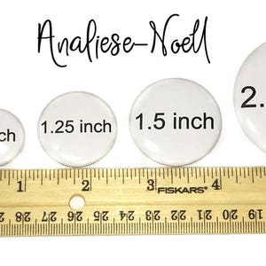 Op de afbeelding: Vier witte ronde knoppen in verschillende maten, met een diameter van 2,5 cm, 3,17 cm, 3,8 cm en 5,7 cm. De knoppen zijn van klein naar groot op een houten liniaal gelegd.