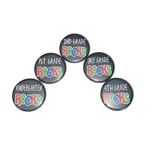 May include: Five round black buttons with white text and colorful lettering that reads "Kindergarten Rocks", "1st Grade Rocks", "2nd Grade Rocks", "3rd Grade Rocks", and "4th Grade Rocks".