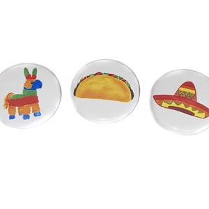 May include: Three round buttons with different Mexican-themed designs. The first button features a colorful pinata donkey. The second button has a yellow taco. The third button shows a red sombrero.