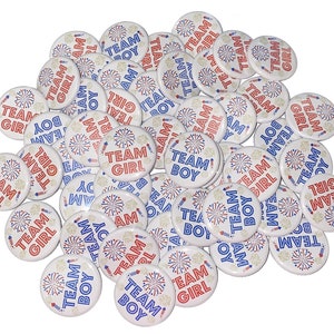May include: A collection of round, white buttons with red and blue text and images. The buttons feature the words "Team Boy" and "Team Girl" with a firework design.