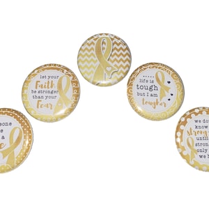 May include: Five gold and white button pins with different inspirational messages and gold ribbon designs. The messages include "someone I love needs a cure", "let your faith be stronger than your fear", "life is tough but I am tougher", and "we don't know how strong we are until being strong is the only choice we have".