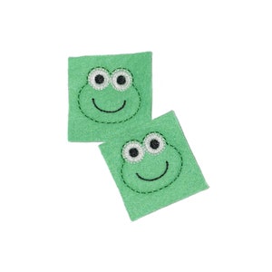 May include: Two green felt squares with embroidered frog faces. Each frog has black eyes, a white pupil, and a black stitched smile.