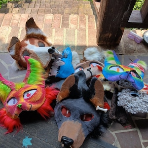 May include: A collection of multiple animal-themed masks featuring various colors and textures. The masks include a brown and white canine, a bright neon pink and green feline, a dark grey wolf with a bone accessory, and a rainbow-colored mask.