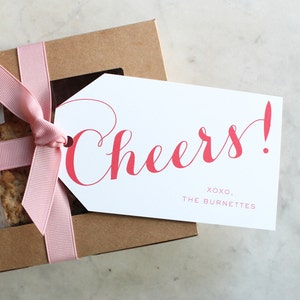 May include: A brown cardboard box with a pink ribbon and a white gift tag. The gift tag has the word "Cheers!" in red lettering and the text "XOXO, THE BURNETTES".