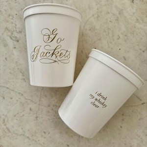 May include: Two white plastic cups with gold lettering. One cup says "Go Jackets" and the other cup says "I drink my whiskey clear."