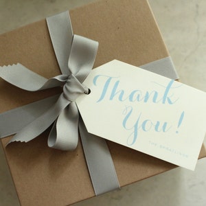May include: A brown gift box with a gray ribbon and bow. A white gift tag with the text "Thank You!" in blue lettering is attached to the box.