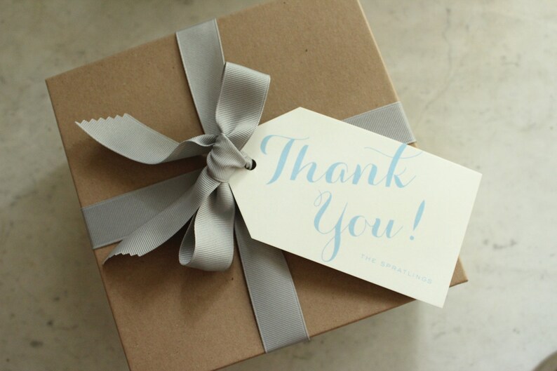 May include: A brown gift box with a gray ribbon and bow. A white gift tag with the text "Thank You!" in blue lettering is attached to the box.