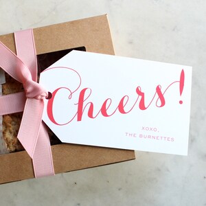 May include: A brown gift box with a pink ribbon and a white gift tag. The tag has the word "Cheers!" in red cursive lettering and the text "XOXO, THE BURNETTES" in smaller red lettering.