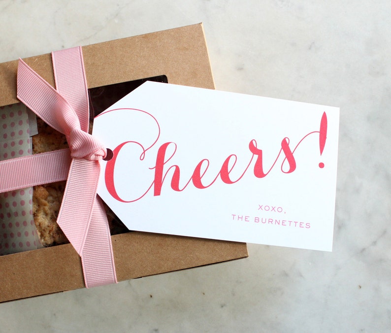 May include: A brown gift box with a pink ribbon and a white gift tag. The tag has the word "Cheers!" in red cursive lettering and the text "XOXO, THE BURNETTES" in smaller red lettering.