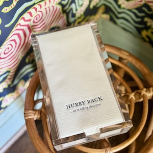 May include: A clear acrylic napkin holder with a white napkin that reads "HURRY BACK...we're talking about you".