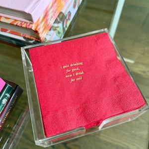May include: A red cocktail napkin with gold text inside a clear acrylic holder. The text reads: "i quit drinking for good, now i drink for evil." The napkin is on a glass table with books in the background.