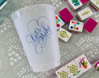 May include: A white plastic cup with the words "Let's  Mahj" written in blue cursive. The cup is sitting on a table with a variety of colorful mahjong tiles.