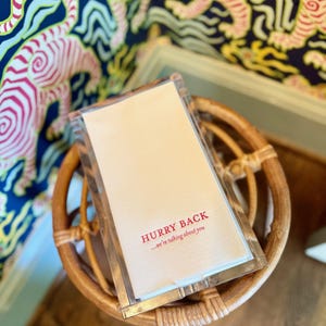 May include: A clear plastic-wrapped stack of white paper napkins with the red text "HURRY BACK ...we're talking about you" sits on a small, round, woven wicker table. The background features a blue and white tiger print.