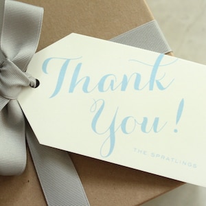May include: A white gift tag with a blue cursive "Thank You!" message and the text "The Spratlings" at the bottom. The tag is tied to a brown gift box with a gray ribbon.