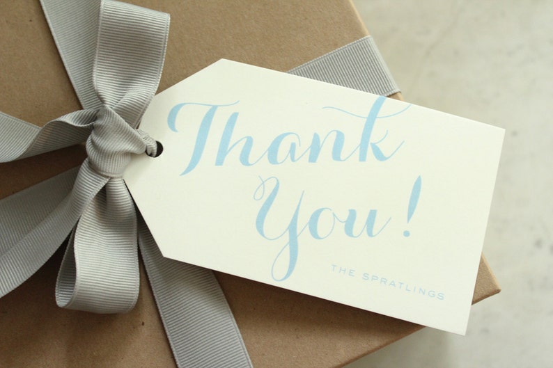 May include: A white gift tag with a blue cursive "Thank You!" message and the text "The Spratlings" at the bottom. The tag is tied to a brown gift box with a gray ribbon.