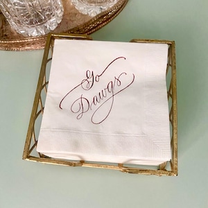May include: A stack of white paper napkins held in a square gold-toned metal holder. The top napkin features the text Go Dawgs written in a dark red cursive script. The napkins have a textured border along the right edge and are square in shape.
