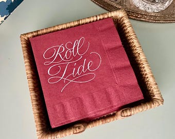 May include: A stack of square, deep red paper napkins featuring the white cursive text Roll Tide, contained within a woven light brown wicker basket. The napkins have a textured border and are positioned on a flat, light-colored surface.