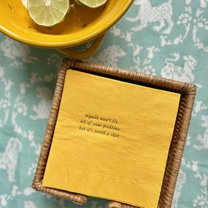 May include: A yellow napkin with the text "Tequila won't fix all of your problems but it's worth a shot" printed on it. The napkin is in a woven wicker napkin holder.