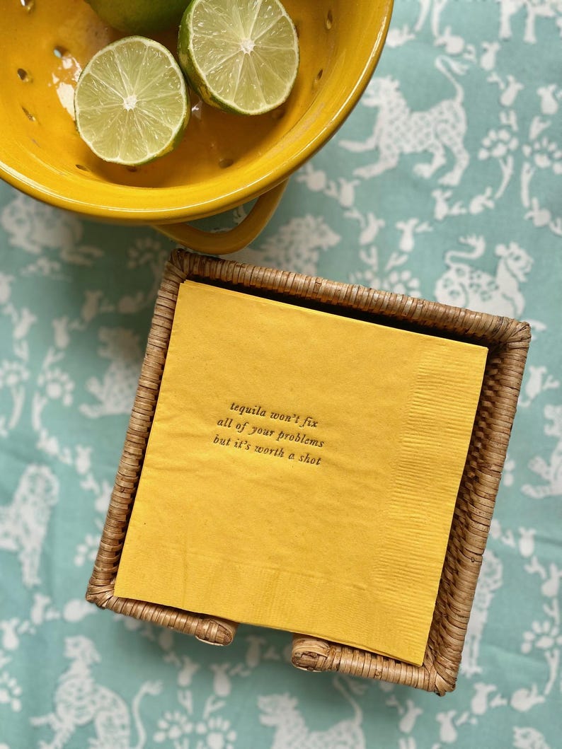 May include: A yellow napkin with the text "Tequila won't fix all of your problems but it's worth a shot" printed on it. The napkin is in a woven wicker napkin holder.