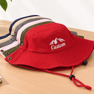 May include: A stack of six bucket hats in red, olive green, light pink, grey, beige, and navy blue. Each hat features a wide brim, metal side eyelets, and a chin strap with a toggle. The red hat displays a white mountain graphic and the word Custom.