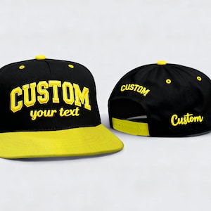 May include: Black and yellow custom snapback hat. The hat features a black crown with the word "CUSTOM" and "your text" in yellow. The brim is yellow, and the hat has a yellow button on top. The back of the hat also has the word "CUSTOM" in yellow.