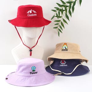 K&ouml;nnte beinhalten: Eine Auswahl an Fischerh&uuml;ten in verschiedenen Farben, darunter Rot, Beige, Marineblau und Lavendel. Jeder Hut hat ein gesticktes Design, wie eine Berglandschaft, eine Blume oder die W&ouml;rter "Adventure" oder "Camping". Die H&uuml;te haben Kinnriemen.